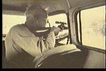 A man pointing a scoped rifle out of his car window at an animal